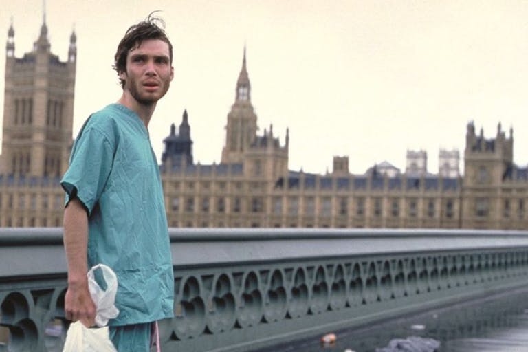 A young skinny white man in hospital scrubs stands on an empty bridge in London with the Parliament building behind him 