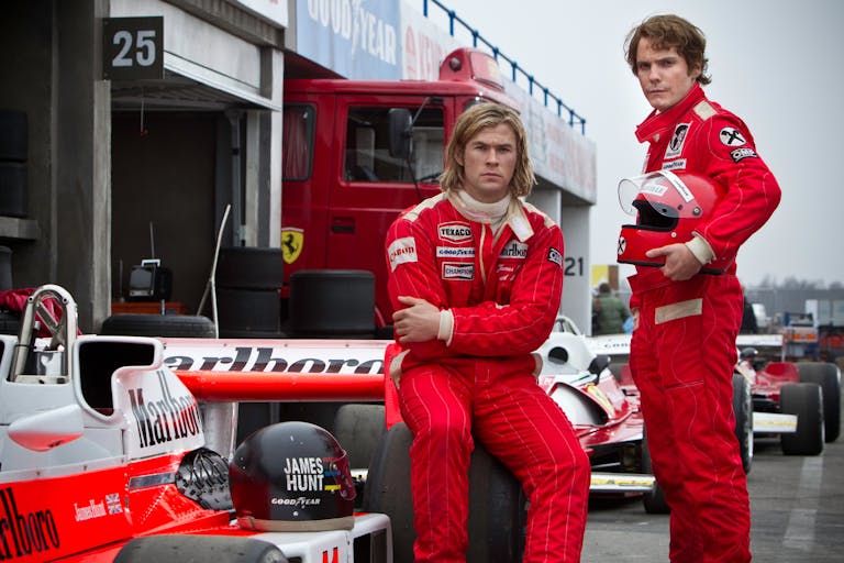 Two men in F1 drivers jumpsuits one sitting, one standing beside a race car, both in red