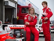 Two men in F1 drivers jumpsuits one sitting, one standing beside a race car, both in red