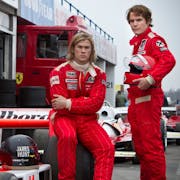 Two men in F1 drivers jumpsuits one sitting, one standing beside a race car, both in red