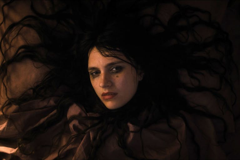 A bedraggled white woman with long black hair splayed out around her, wearing black clothing and smudged black eye make up, looking serious
