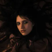 A bedraggled white woman with long black hair splayed out around her, wearing black clothing and smudged black eye make up, looking serious