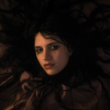 A bedraggled white woman with long black hair splayed out around her, wearing black clothing and smudged black eye make up, looking serious 