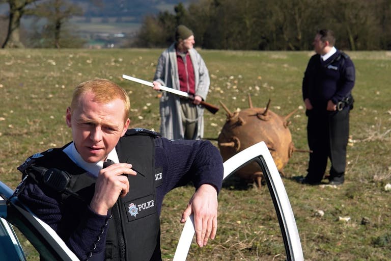Two white male police officers standing in a field with a white bedraggled man holding a shotgun next to a large circular ancient undetonated bomb