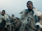 A Black man flanked by two brown women and a Black man in a cold wilderness wearing light coloured animal furs