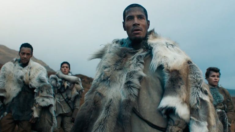 A Black man flanked by two brown women and a Black man in a cold wilderness wearing light coloured animal furs