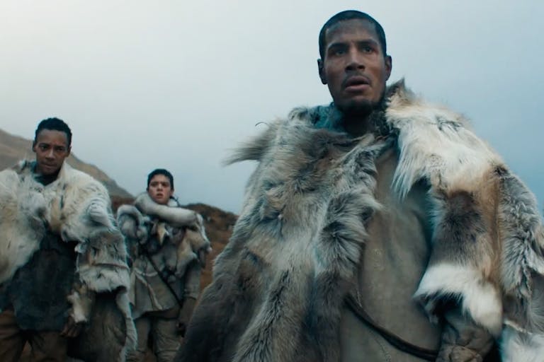 A Black man flanked by two brown women and a Black man in a cold wilderness wearing light coloured animal furs