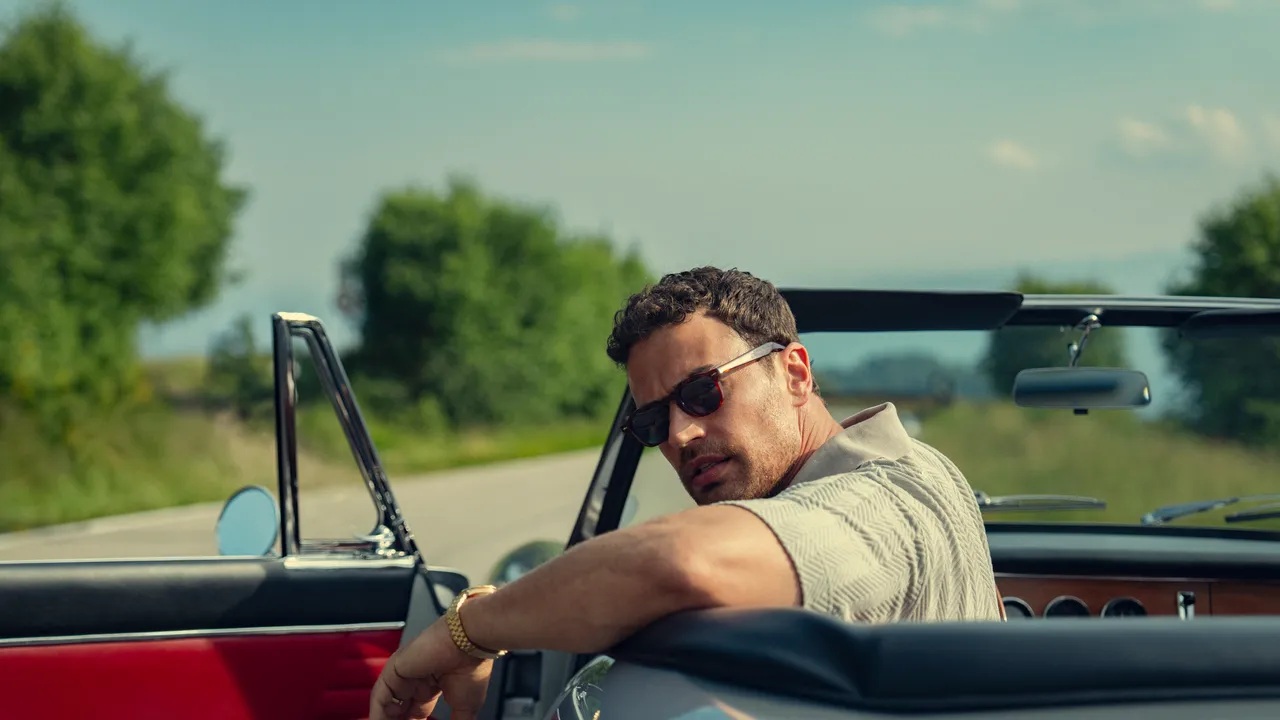 A middle-aged tanned-skin man wearing sunglasses, resting his arm on the side of his roofless car and looking back