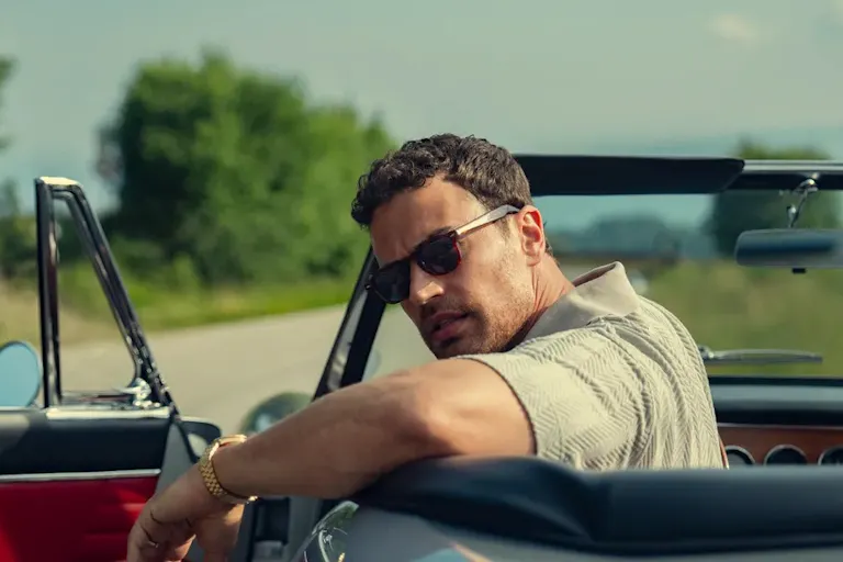 A middle-aged tanned-skin man wearing sunglasses, resting his arm on the side of his roofless car and looking back