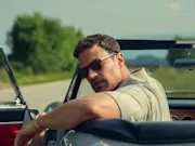 A middle-aged tanned-skin man wearing sunglasses, resting his arm on the side of his roofless car and looking back