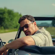 A middle-aged tanned-skin man wearing sunglasses, resting his arm on the side of his roofless car and looking back