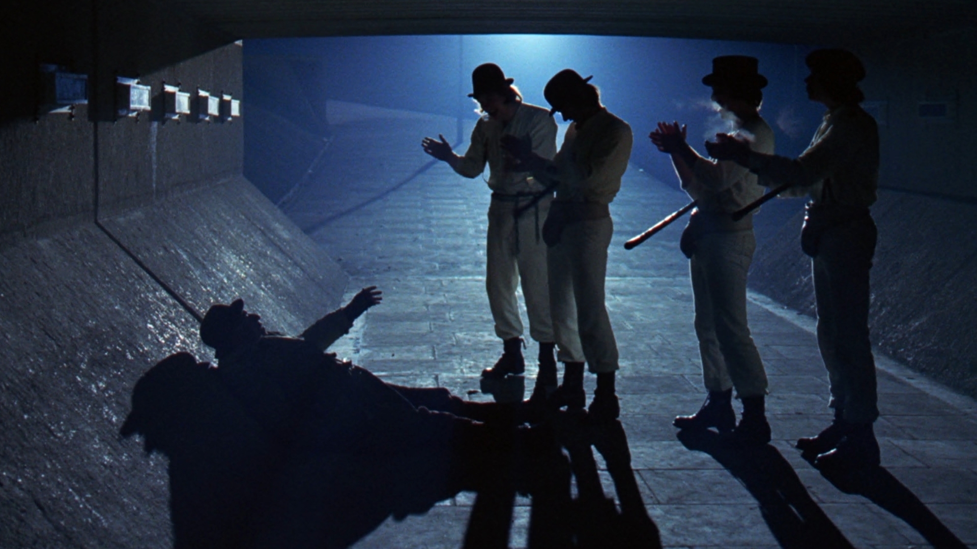 4 men in matching white suits and bowler hats, standing over a shadow-covered man under a bridge at night time