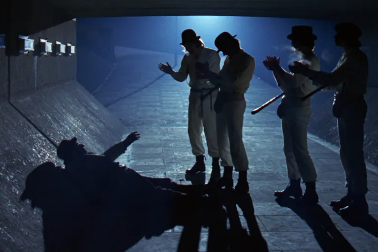4 men in matching white suits and bowler hats, standing over a shadow-covered man under a bridge at night time