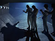 4 men in matching white suits and bowler hats, standing over a shadow-covered man under a bridge at night time