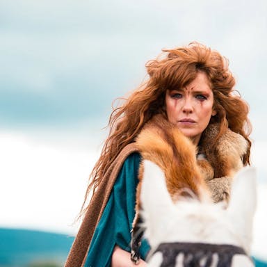 Woman with long red hair stands outside, wearing a fur outfit and tribal make-up. 