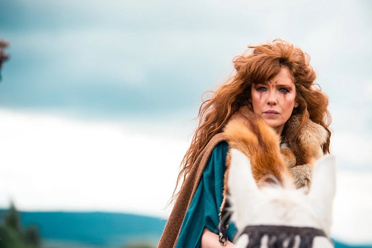 Woman with long red hair stands outside, wearing a fur outfit and tribal make-up.