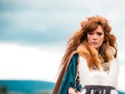 Woman with long red hair stands outside, wearing a fur outfit and tribal make-up.