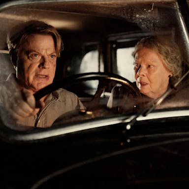 A middle aged male character looking determined and an old woman sit in an old car looking shocked