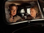A middle aged male character looking determined and an old woman sit in an old car looking shocked