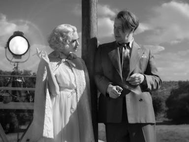Black and white still of a white blonde woman dressed in a white dress and cape smoking a cigarette, lent against a pole next to a white man in a dark suit