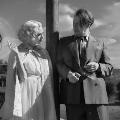 Black and white still of a white blonde woman dressed in a white dress and cape smoking a cigarette, lent against a pole next to a white man in a dark suit