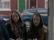 Two young white girls wearing loose head scarves standing in a doorway handing out pamphlets