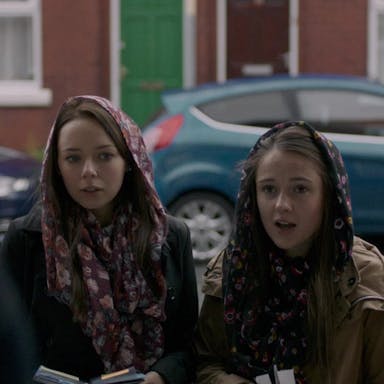Two young white girls wearing loose head scarves standing in a doorway handing out pamphlets