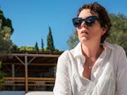 Middle aged woman with short dark hair, wearing dark sunglasses and a white shirt, looks into the distance against the backdrop of a blue sky and green lush trees.