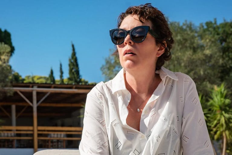 Middle aged woman with short dark hair, wearing dark sunglasses and a white shirt, looks into the distance against the backdrop of a blue sky and green lush trees.