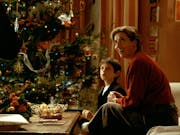 Smiling woman with short blonde hair sits with a young boy on a sofa in front of a decorated Christmas tree.