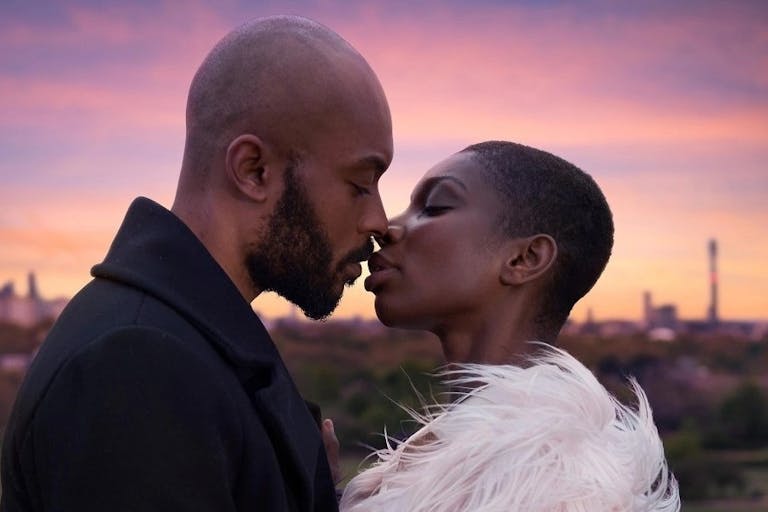 A young black woman and man kiss in front of a sunset view of  the London sky.