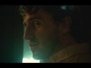 A young white man with angular features looks to the side in dark lighting