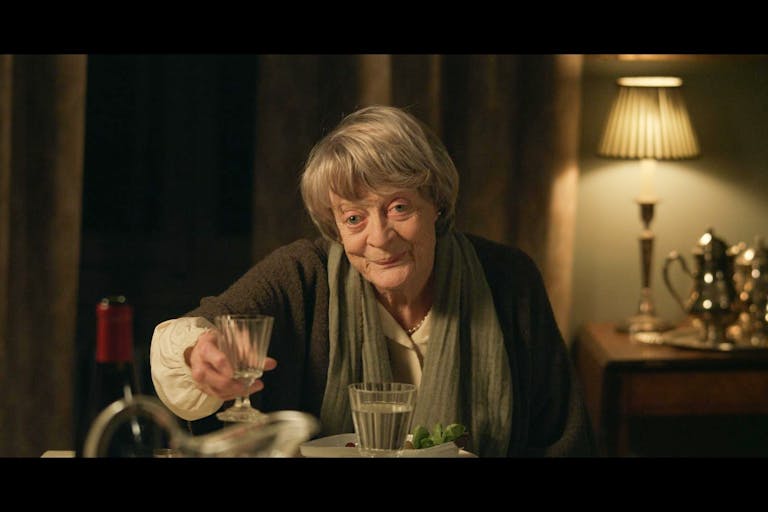 An old woman sits at a table at night, toasting a glass