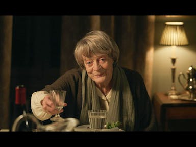 An old woman sits at a table at night, toasting a glass