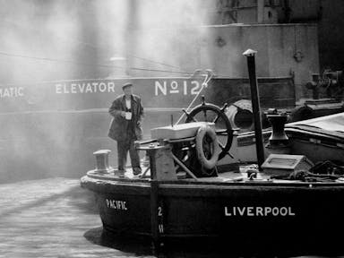 A black and white still of a middle aged man stood at the helm of an old cargo boat