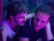 A young white man with his arm around a middle aged white man in a club setting with fluorescent purple light on them