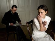 Young woman in white dress looks anguished sitting on a wooden chair, mustached man in old-fashioned suit sits behind her, writing in a notebook.