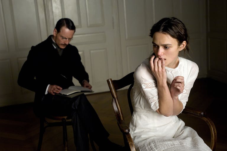 Young woman in white dress looks anguished sitting on a wooden chair, mustached man in old-fashioned suit sits behind her, writing in a notebook.