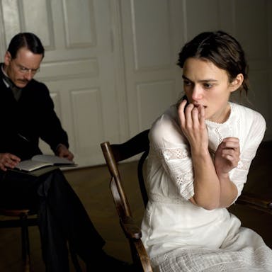 Young woman in white dress looks anguished sitting on a wooden chair, mustached man in old-fashioned suit sits behind her, writing in a notebook. 