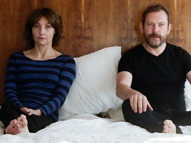 A woman and a man sitting in bed on top of white covers, wearing dark clothes looking ahead