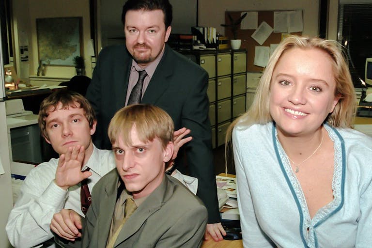 Three white men looking grumpy and one white woman smiling in dated office attire in a boring office setting