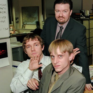 Three white men looking grumpy and one white woman smiling in dated office attire in a boring office setting