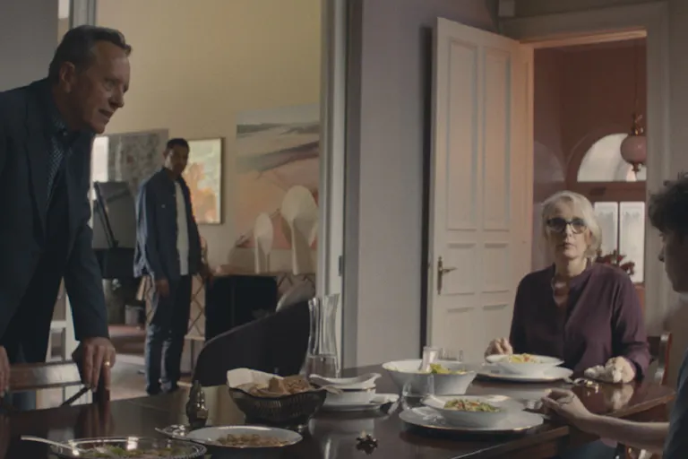 A man stands behind a dinner chair looking at a young boy sat at the table along with a woman eating food. A young man stands looking on in the background.