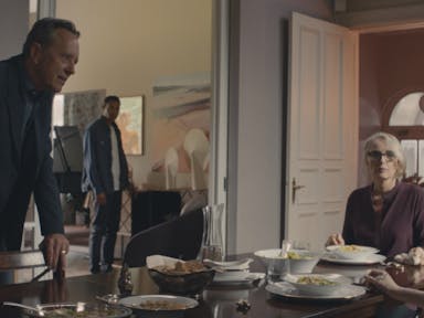 A man stands behind a dinner chair looking at a young boy sat at the table along with a woman eating food. A young man stands looking on in the background. 