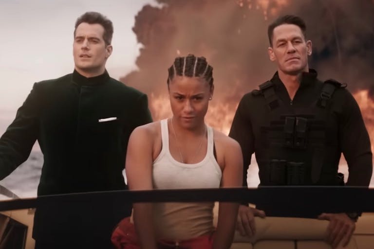 A Black woman in a white vest top and two white men in black suit and black combat clothes riding in a speed boat