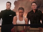A Black woman in a white vest top and two white men in black suit and black combat clothes riding in a speed boat