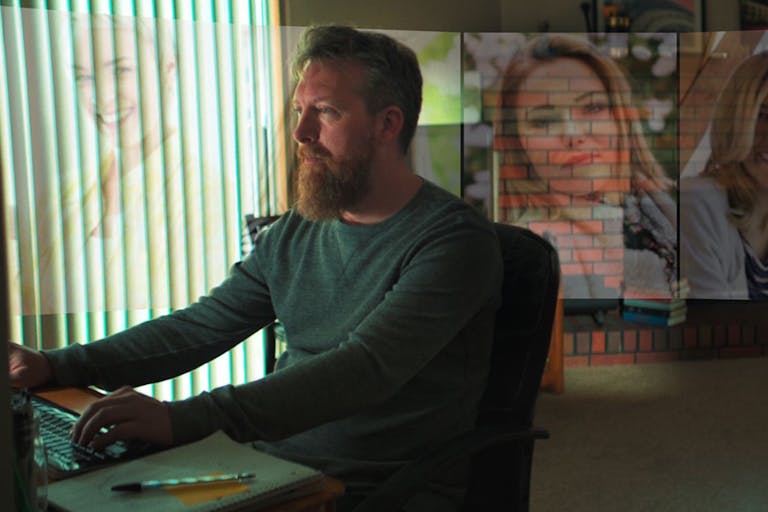 A white man sits at his computer with projected images of white woman around him