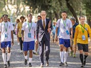 A line of young Black footballers in England kit walking with an older white man in a suit on a sunny tree lined path