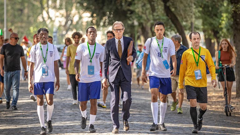 A line of young Black footballers in England kit walking with an older white man in a suit on a sunny tree lined path