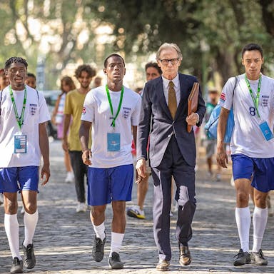 A line of young Black footballers in England kit walking with an older white man in a suit on a sunny tree lined path